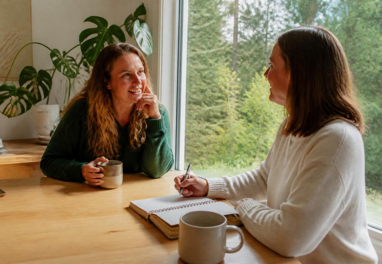 Two people talking over coffee and notes at a sunlit table overlooking British Columbia forest